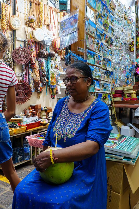 Femme en robe bleue assise qui se désaltère en buvant du lait de coco avec une paille, dans une coursive du marché de Port-louis. Île Maurice, 2026.